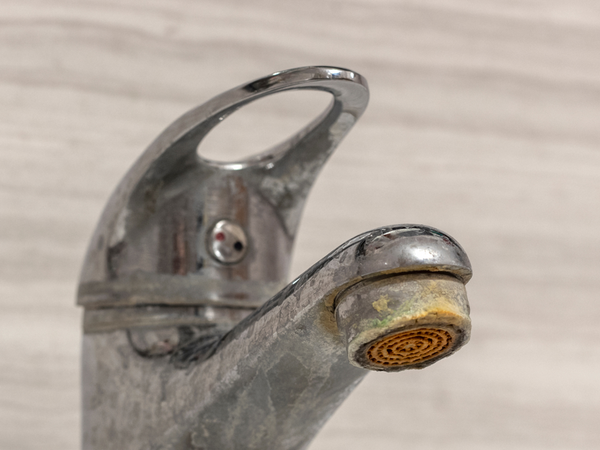 A chrome bathroom faucet tap heavily coated with dull white limescale stains and yellowish hard water mineral crust buildup around the aerator nozzle