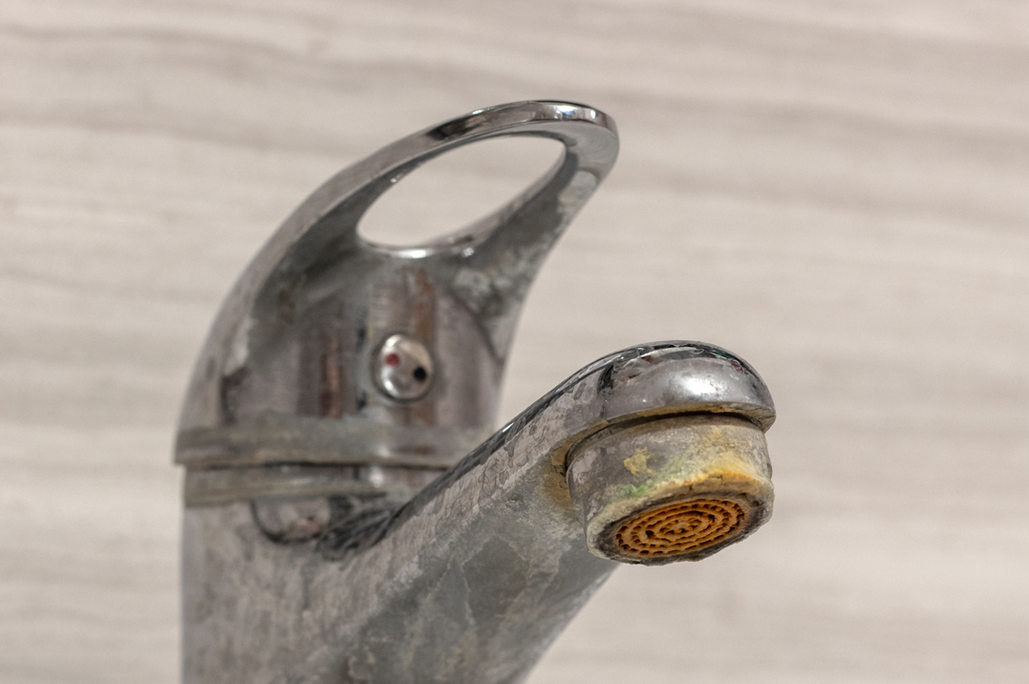 A chrome bathroom faucet tap heavily coated with dull white limescale stains and yellowish hard water mineral crust buildup around the aerator nozzle