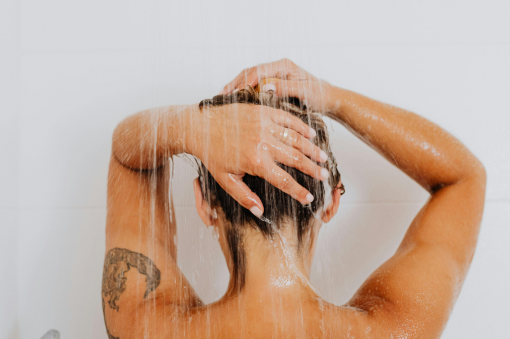 Back view of a woman washing her hair under a bathroom shower stream that contains hidden chlorine minerals and hard water impurities