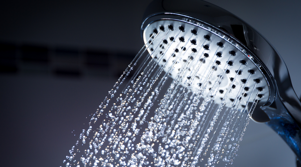Close up of a chrome regular shower head spraying multiple streams of unconditioned bathroom water against a dark tiled wall background
