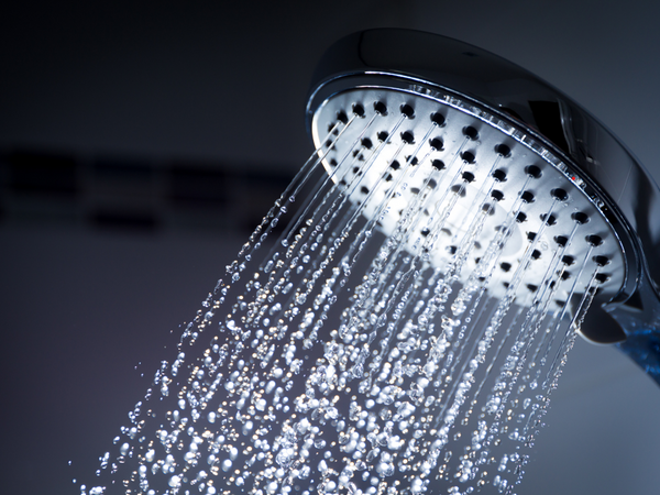 Close up of a chrome regular shower head spraying multiple streams of unconditioned bathroom water against a dark tiled wall background