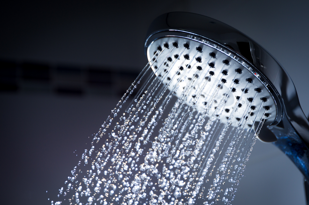 Close up of a chrome regular shower head spraying multiple streams of unconditioned bathroom water against a dark tiled wall background